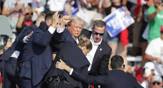 Donald Trump ferito, col sangue che gli cola dall'orecchio, sostenuto dagli agenti, col pugno alzato